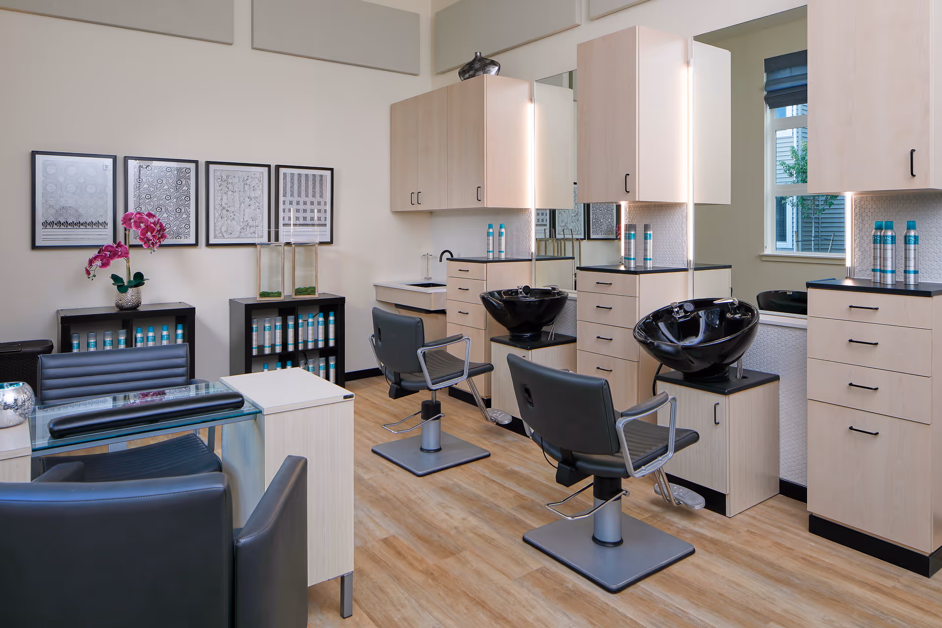 Interior of a modern salon area with two black salon chairs facing black wash basins. Light wood cabinetry with mirrors and shelves holding hair products. A glass table with black chairs and decorative framed artwork on the wall. Light wood flooring and a window with a blue shade.