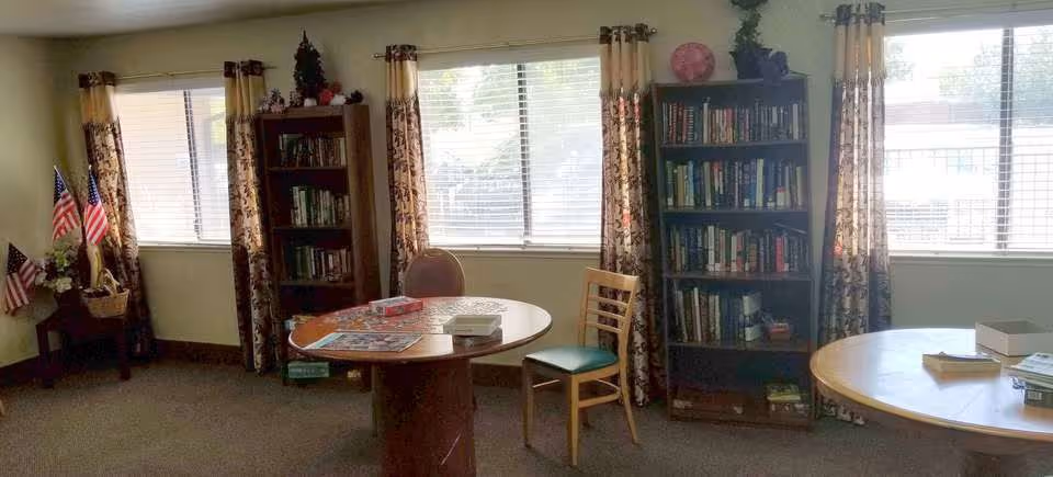 A cozy room with two round wooden tables, one with a chair and board games on top. There are two tall bookshelves filled with books against the wall with windows covered by floral curtains. A small corner table holds a basket with American flags and a decorative eagle statue.