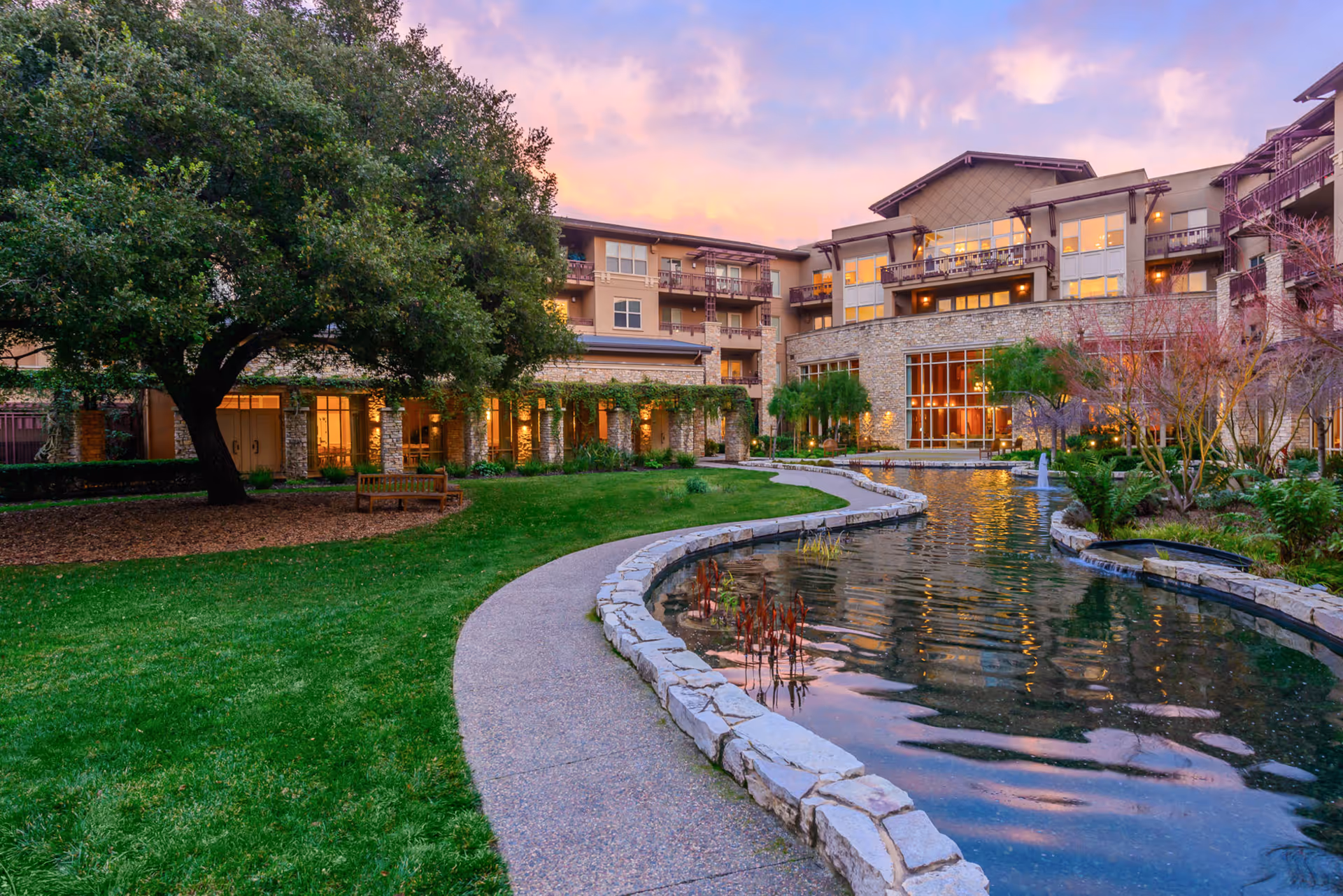Courtyard with a curved stone-lined pond, pathway, and large tree in front of a multi-story senior living building at sunset.