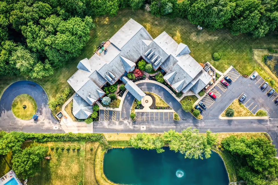 Aerial view of the Saint Therese of Westlake facility showing a large building with a gray roof surrounded by green trees and lawns. There is a curved driveway and parking lot with several cars parked. A pond with a small fountain is visible in the lower part of the image.