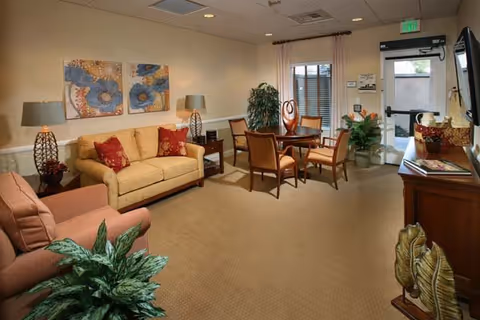 Comfortable lounge with a sofa, armchairs, a small round dining table, decorative plants and wall art near a glass exit door.