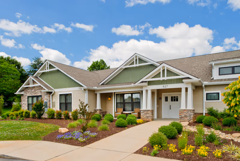 Front exterior view of a single-story senior living facility building with a beige and green facade, stone accents, a covered entrance, and well-maintained landscaping with bushes and flowers under a partly cloudy blue sky.
