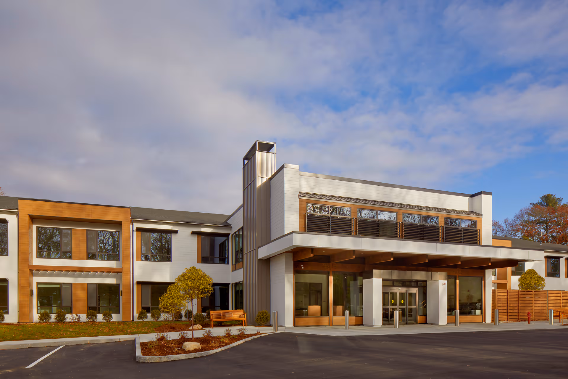 Exterior view of The Cordwainer senior living facility showing a modern two-story building with large windows, wood and white paneling, a covered entrance, landscaped shrubs, and a parking area in front under a partly cloudy sky.