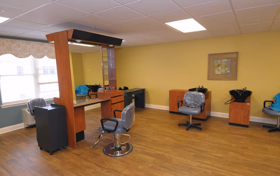 Interior view of a hair salon area with wooden flooring, yellow walls, and large windows with blinds. The room contains salon chairs, hair washing stations with sinks, a large mirror with a wooden frame, and a framed artwork on the wall.