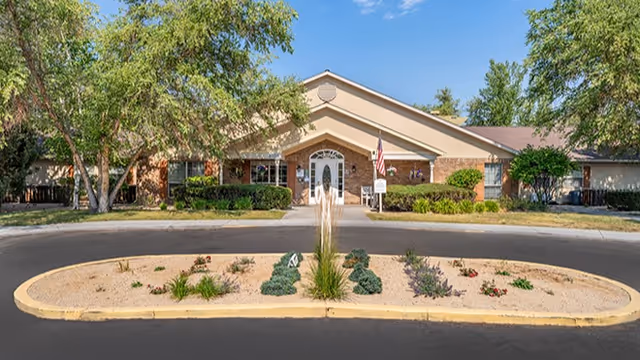 Front exterior view of a single-story senior living facility building with a peaked roof, brick facade, and white double doors. There is a circular driveway with a landscaped island in the center featuring plants and shrubs. Trees and bushes surround the building under a clear blue sky.