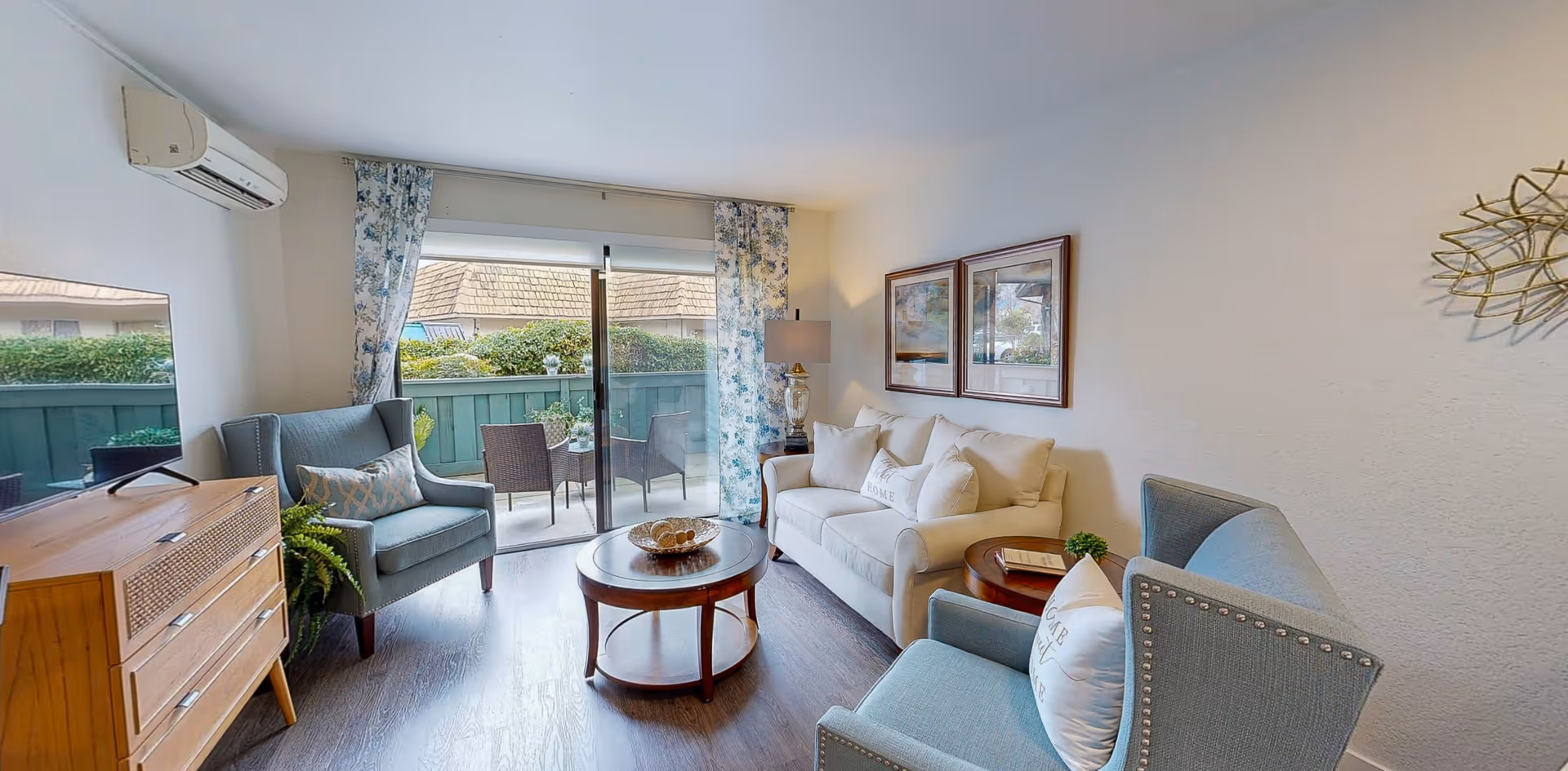 A cozy living room with a white sofa, two blue armchairs, a round wooden coffee table, and a wooden cabinet with a TV on top. The room has a sliding glass door leading to a small outdoor patio with chairs and a table. The walls are decorated with framed pictures and a decorative wall piece. Light blue floral curtains frame the glass door.