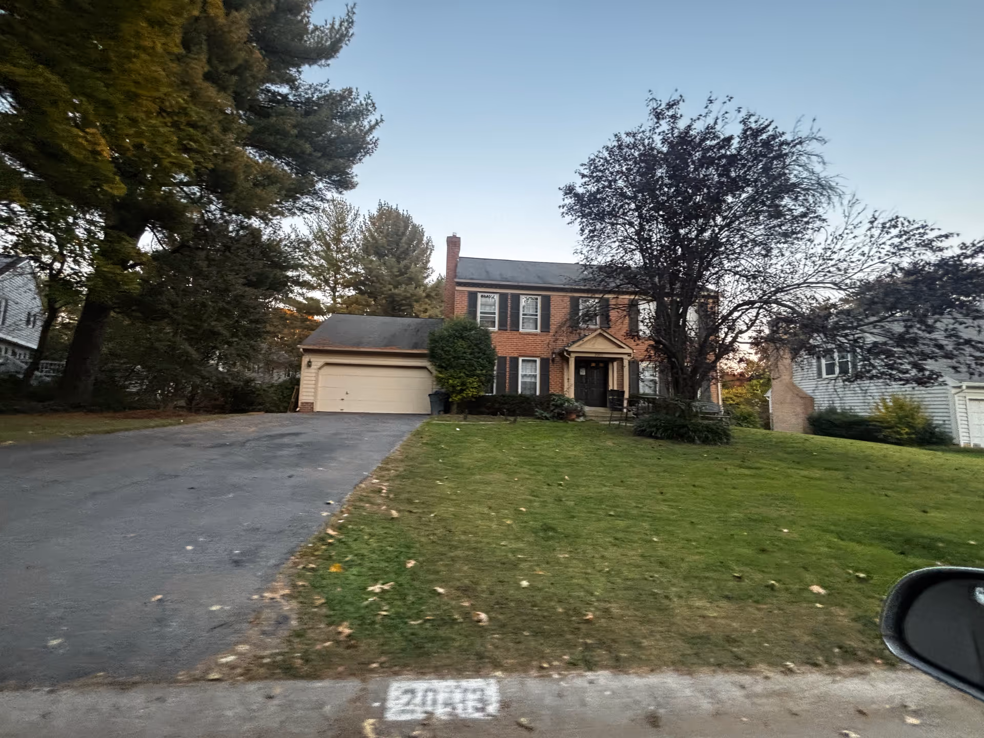 A two-story brick house with black shutters and a black front door, featuring a driveway leading to a two-car garage. The house is surrounded by a grassy lawn with trees on both sides and a clear sky in the background.