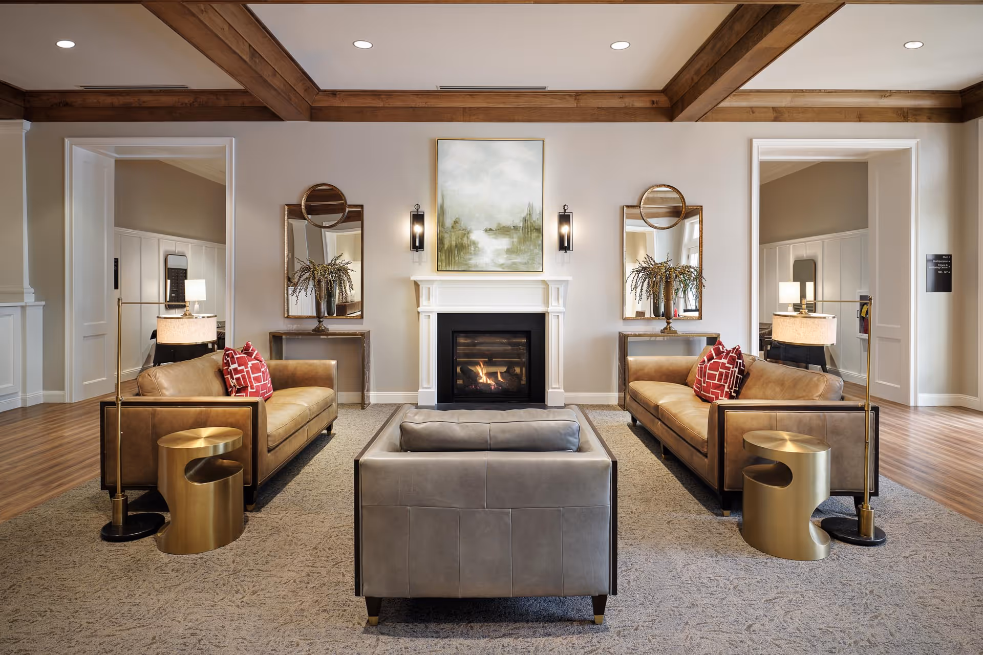 A cozy living room area with a central fireplace, flanked by two brown leather sofas with red patterned pillows, a gray armchair facing the fireplace, gold side tables, floor lamps, and decorative mirrors and plants on console tables against the wall.