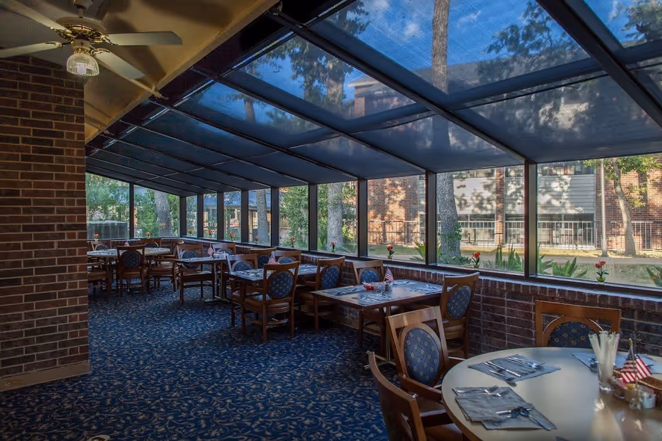 Dining area with multiple tables and chairs arranged neatly. The room has large windows with a view of trees and a brick building outside. The ceiling is slanted with a fan and the floor is carpeted with a patterned design. Tables are set with placemats, utensils, and small American flags.