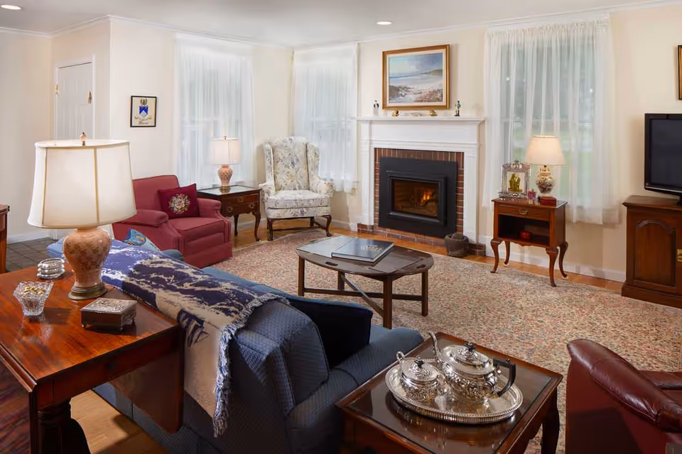 A cozy living room with a lit fireplace, featuring a blue sofa with a patterned throw, a red armchair, a floral wingback chair, wooden side tables with lamps, a coffee table, a large patterned rug, and a television on a wooden stand. The room has cream-colored walls and sheer white curtains on the windows.