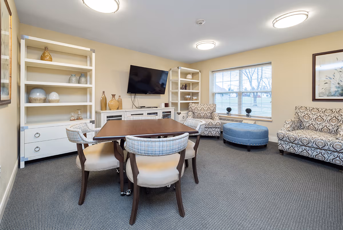 A cozy living room area with a wooden table surrounded by four upholstered chairs in the center. There are two patterned armchairs and a blue round ottoman near a window with blinds. Two white shelving units with decorative vases and a wall-mounted flat-screen TV are visible. The room has beige walls, carpeted floor, and ceiling lights.