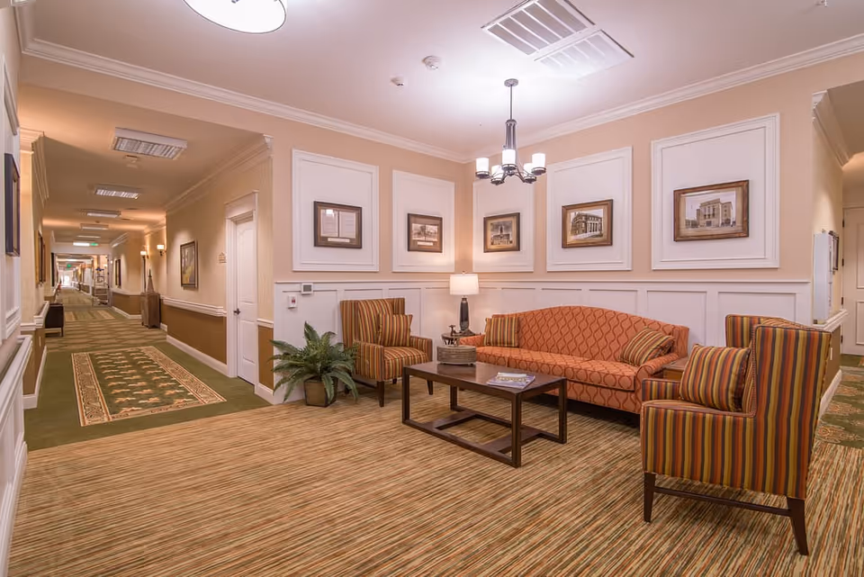 A cozy seating area in a senior living facility hallway with an orange patterned sofa, two striped armchairs, a wooden coffee table with a magazine, a table lamp, framed pictures on the wall, and a green potted plant. The hallway has beige walls, carpeted floors with a green runner, and ceiling lights.