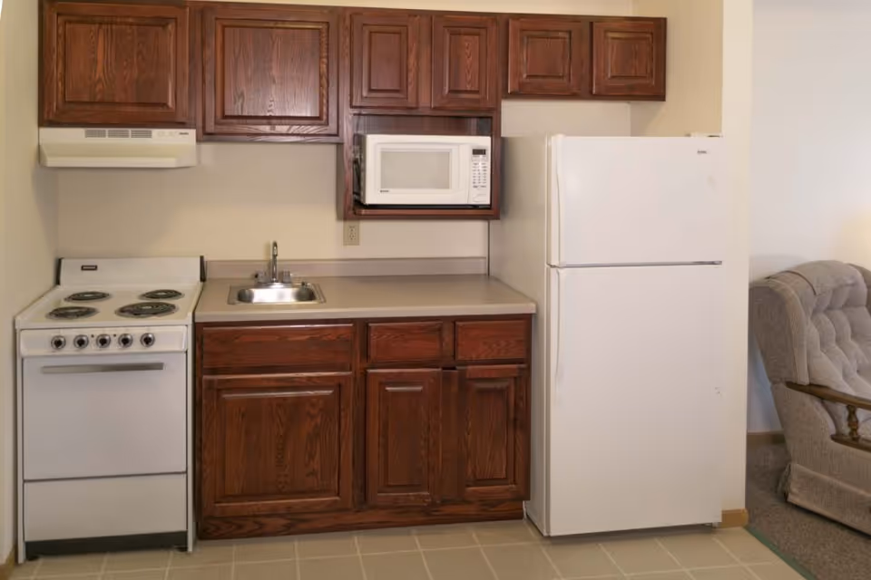 Small kitchen area with a white electric stove, a stainless steel sink, a white microwave mounted above the counter, dark wooden cabinets, and a white refrigerator. A beige upholstered armchair is partially visible to the right.