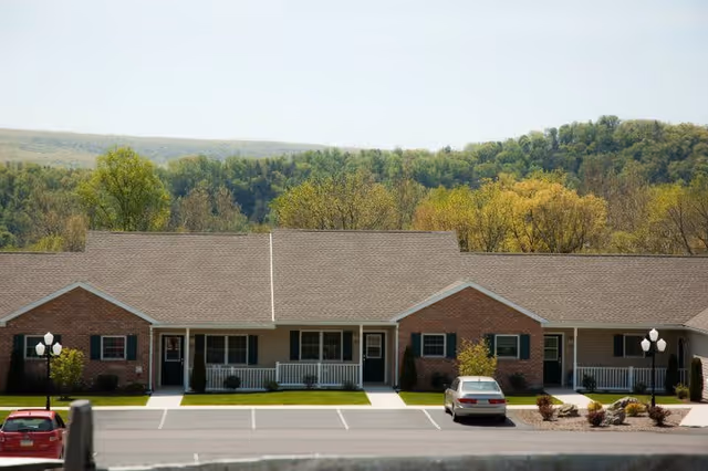 Exterior view of a single-story senior living facility building with brick and siding facade, multiple entrances, a parking lot with cars, and a backdrop of green trees and hills under a clear sky.