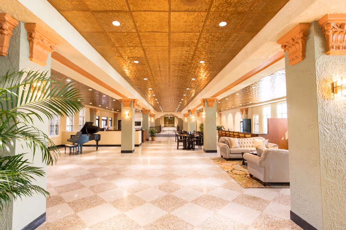 Spacious, ornate interior lounge with sofas, a grand piano, tables, columns and a decorative gold ceiling.
