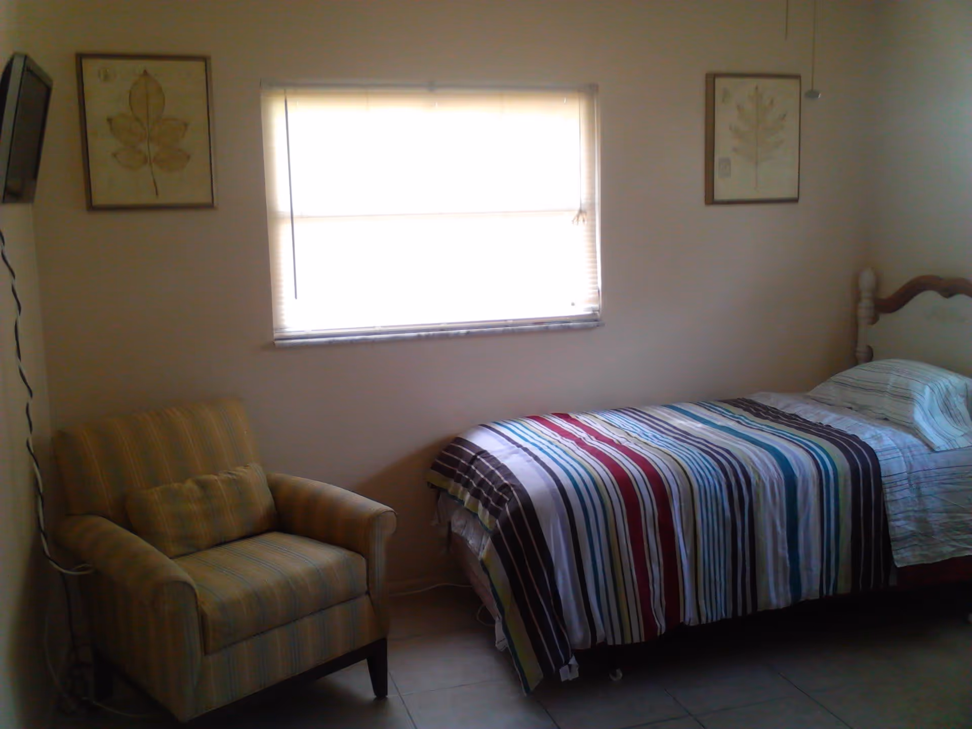 Small bedroom with a single bed dressed in striped bedding, an armchair, and a window between framed prints.
