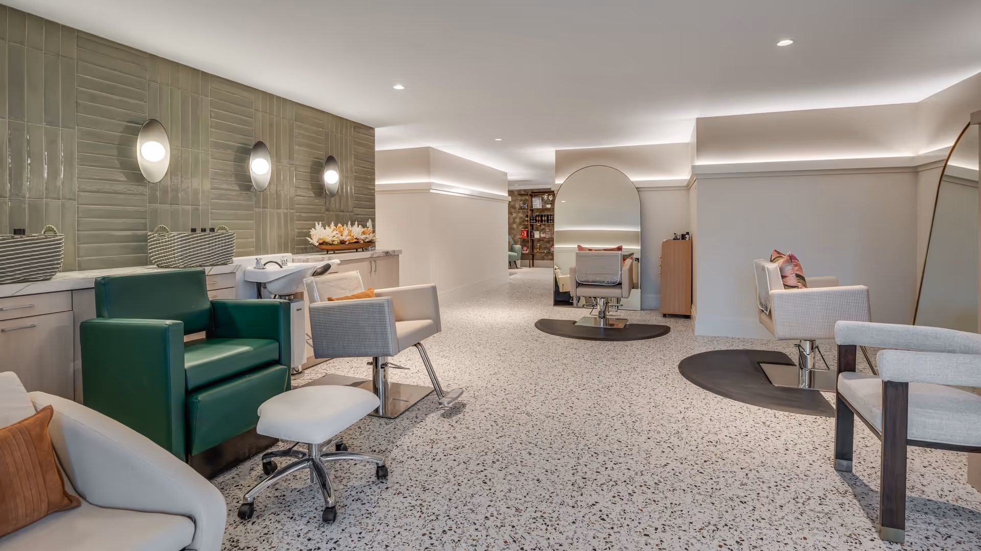 Interior view of a modern salon area with multiple styling chairs, large mirrors, and a green shampoo chair. The space features terrazzo flooring, soft lighting, and a neutral color palette with decorative pillows and wall-mounted lights.