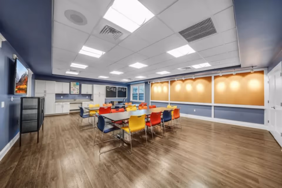 Bright community dining/activity room with long tables, colorful chairs, a kitchenette along the back wall, and large cork boards on one side.