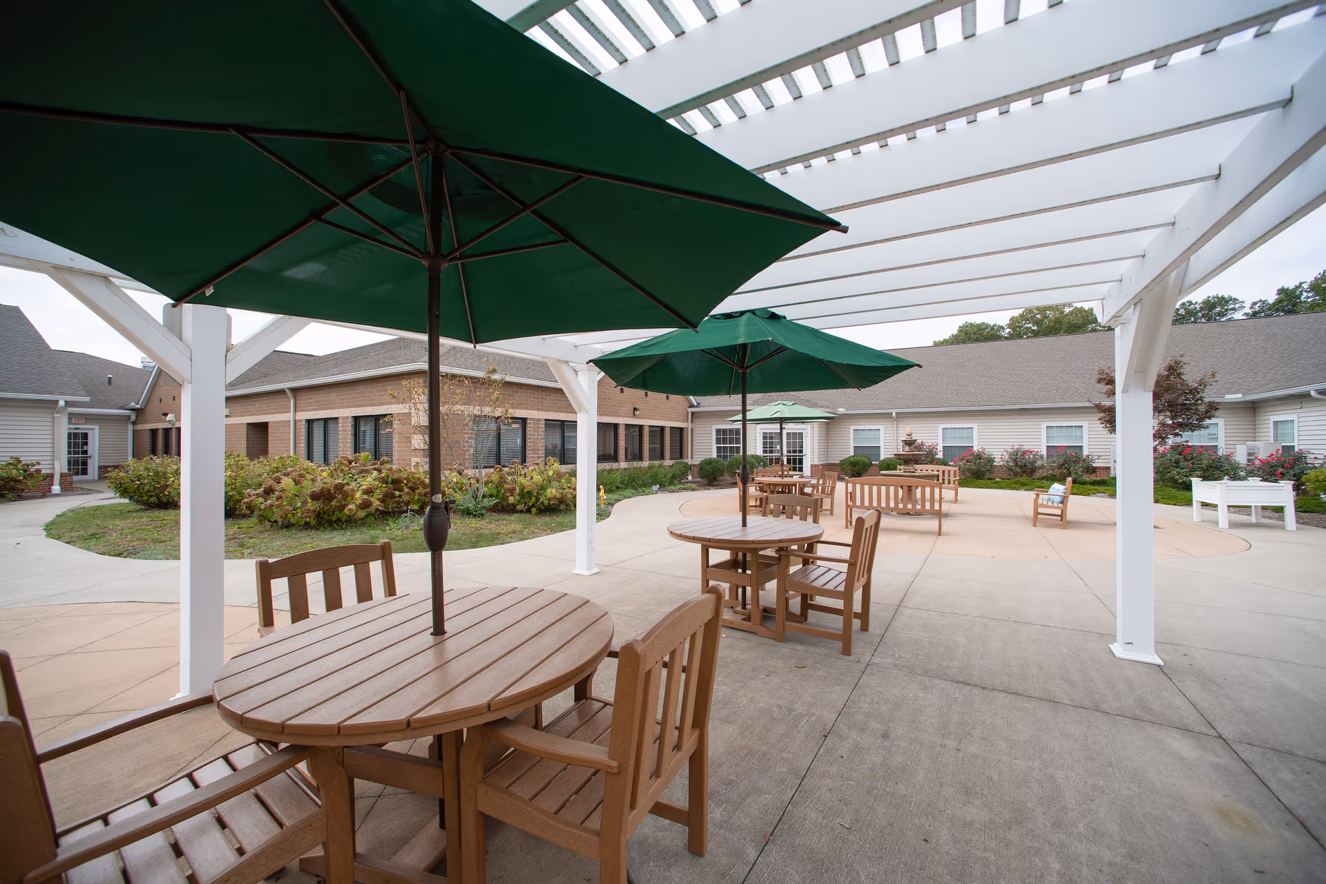 Outdoor patio area at Danbury Massillon with wooden tables and chairs under green umbrellas, surrounded by a white pergola. The patio is paved and has landscaped bushes and flowers around the edges, with a building in the background.