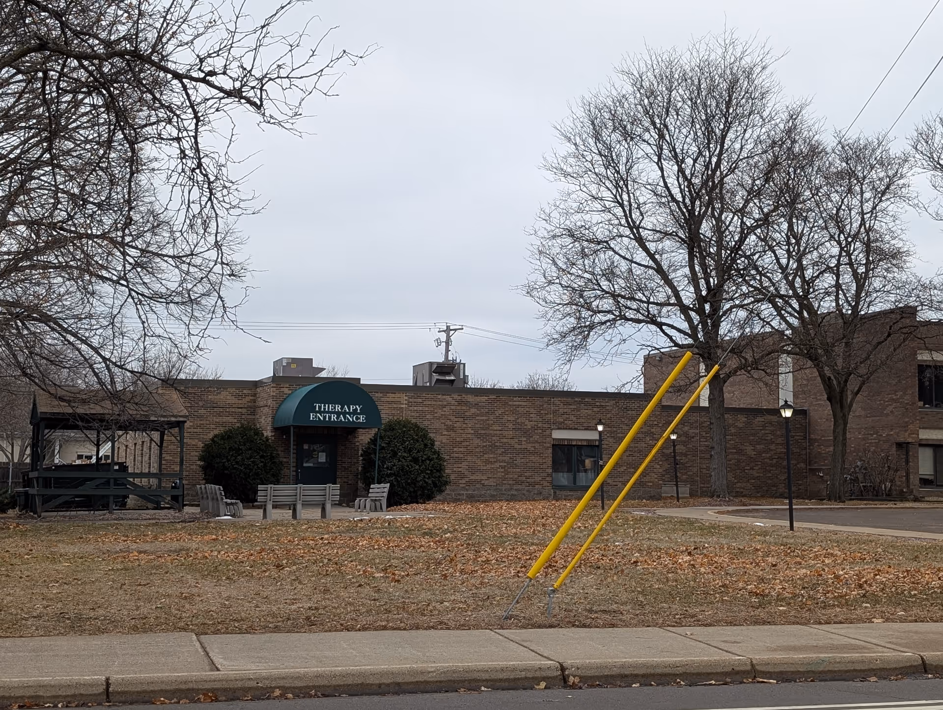 Exterior view of a brick building with a green awning labeled 'THERAPY ENTRANCE'. There are leafless trees, a small gazebo with picnic tables, benches, and a sidewalk in the foreground. The sky is overcast.