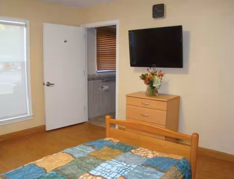 A furnished senior living bedroom with bed, dresser, wall-mounted TV, and an open bathroom door.
