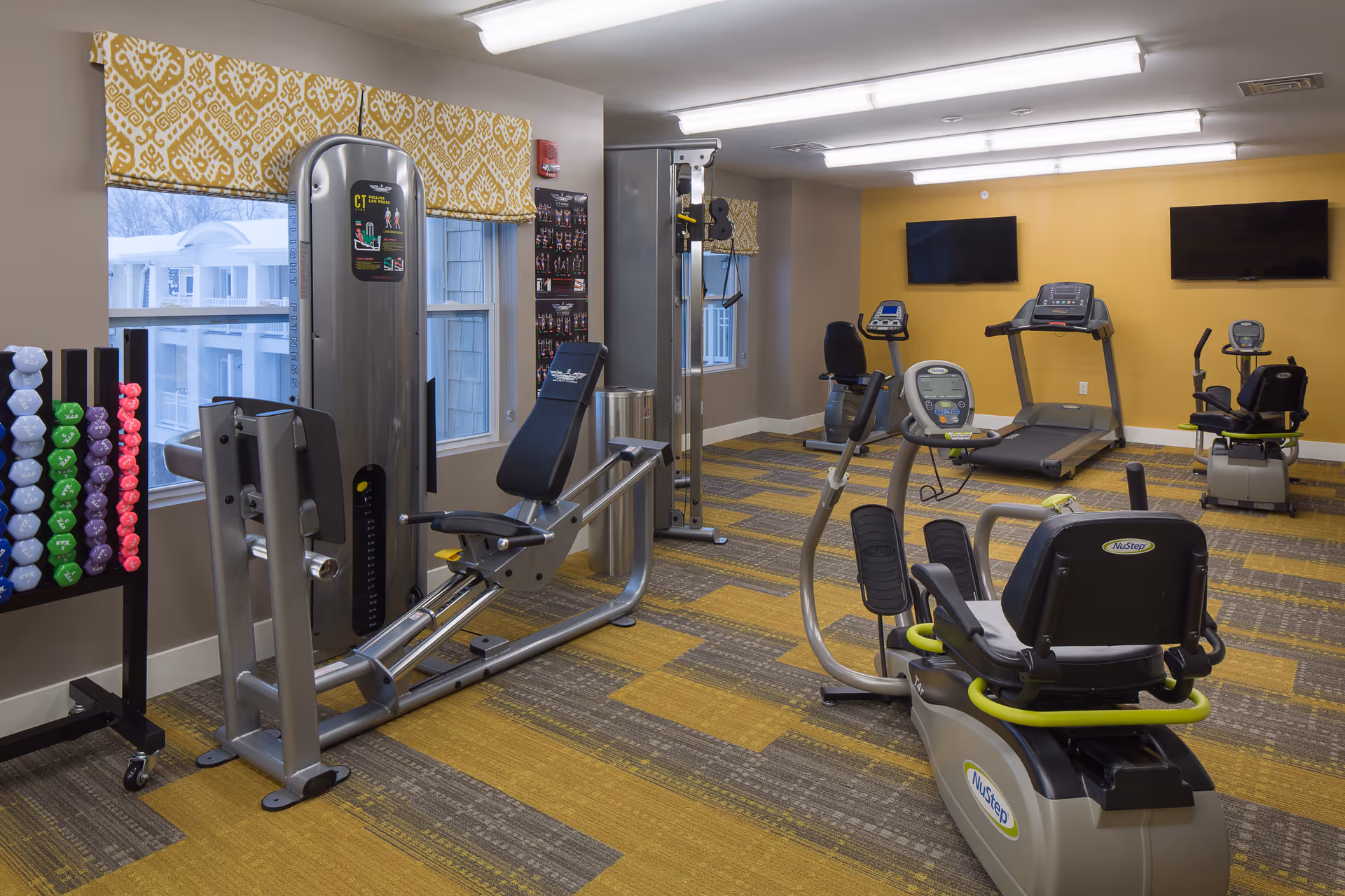 A senior living facility fitness room with various exercise equipment including a recumbent bike, treadmill, weight machine, and a rack of colorful dumbbells. The room has yellow and gray patterned carpet, beige walls, two windows with yellow patterned valances, and two wall-mounted flat-screen TVs on a yellow accent wall.