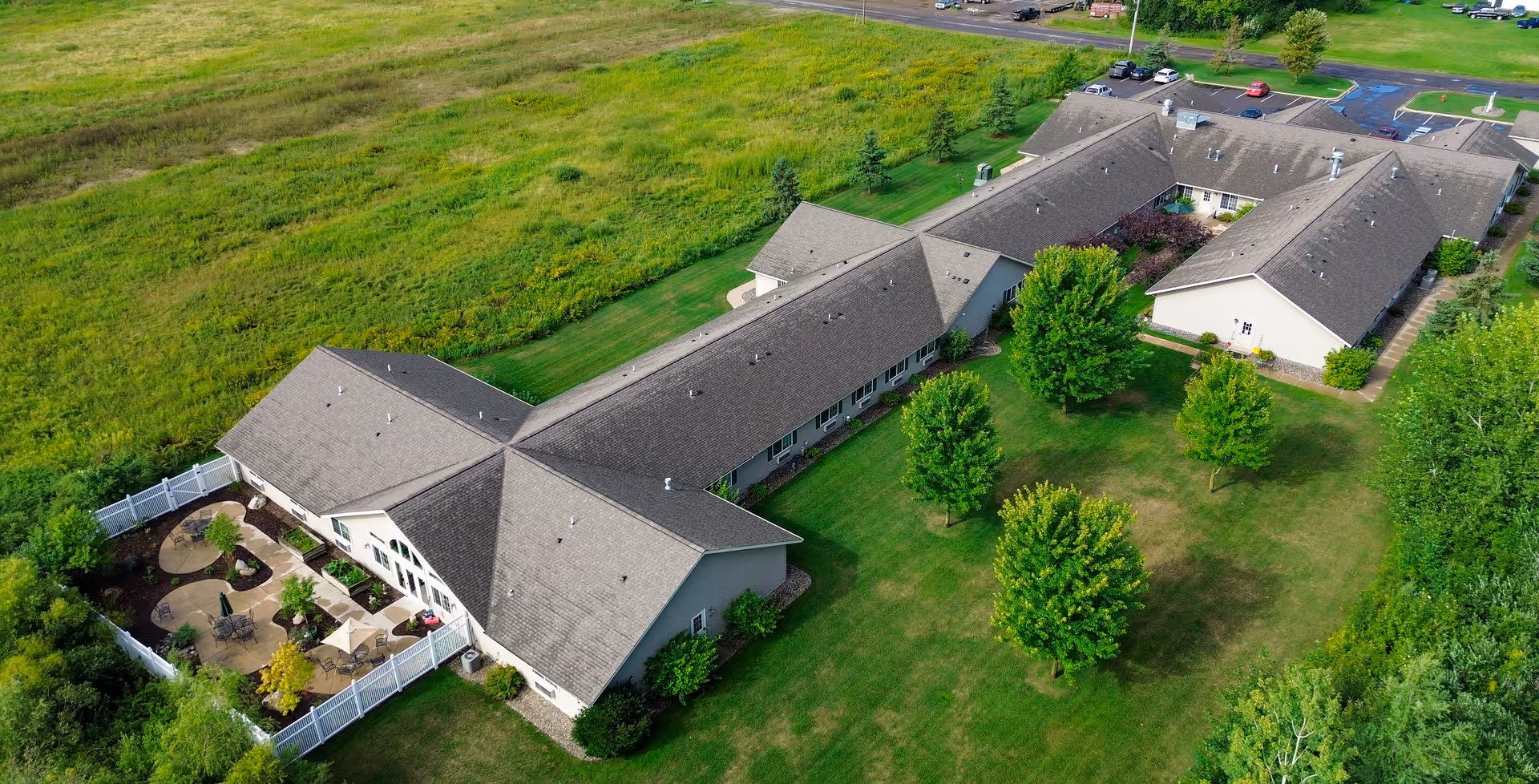 Aerial view of a senior living facility named Golden Horizons of Aitkin, showing a large building with multiple wings surrounded by green lawns, trees, and a fenced patio area with outdoor seating. There is a parking lot visible in the background.