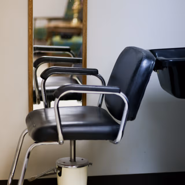 A black salon chair with metal armrests positioned in front of a mirror and next to a black wash basin, inside a room with white walls.