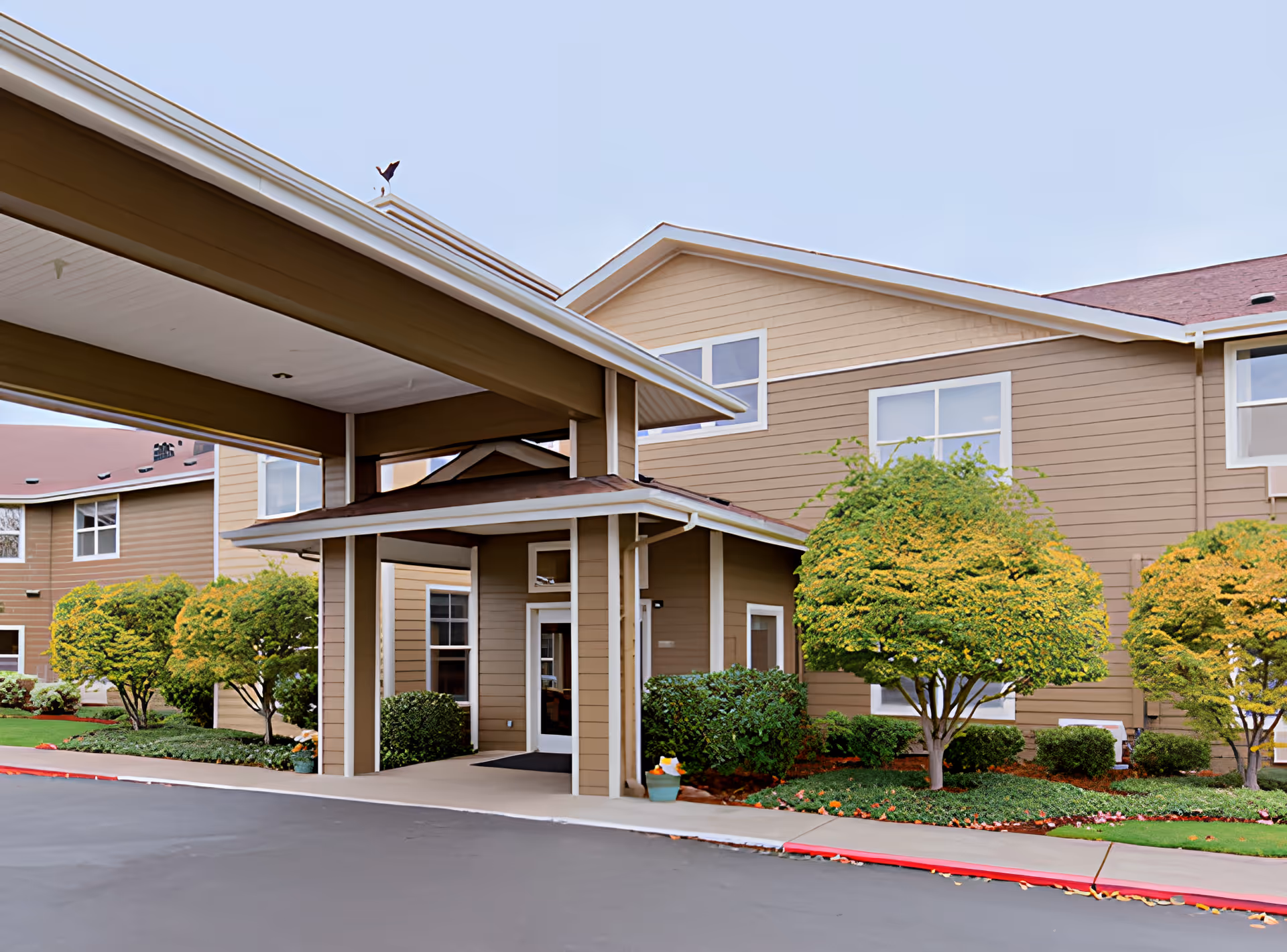 Exterior view of Heron Pointe Senior Living building entrance with a covered drop-off area, beige siding, multiple windows, and well-maintained landscaping including trimmed bushes and small trees.