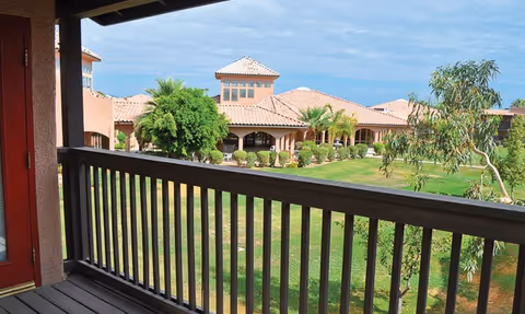 View from a shaded balcony overlooking a grassy courtyard with palm trees and Mediterranean-style senior living buildings.