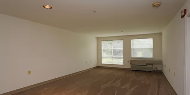 Empty room with beige carpet and white walls featuring two large windows letting in natural light. There is a heating or air conditioning unit below the windows and ceiling lights above.