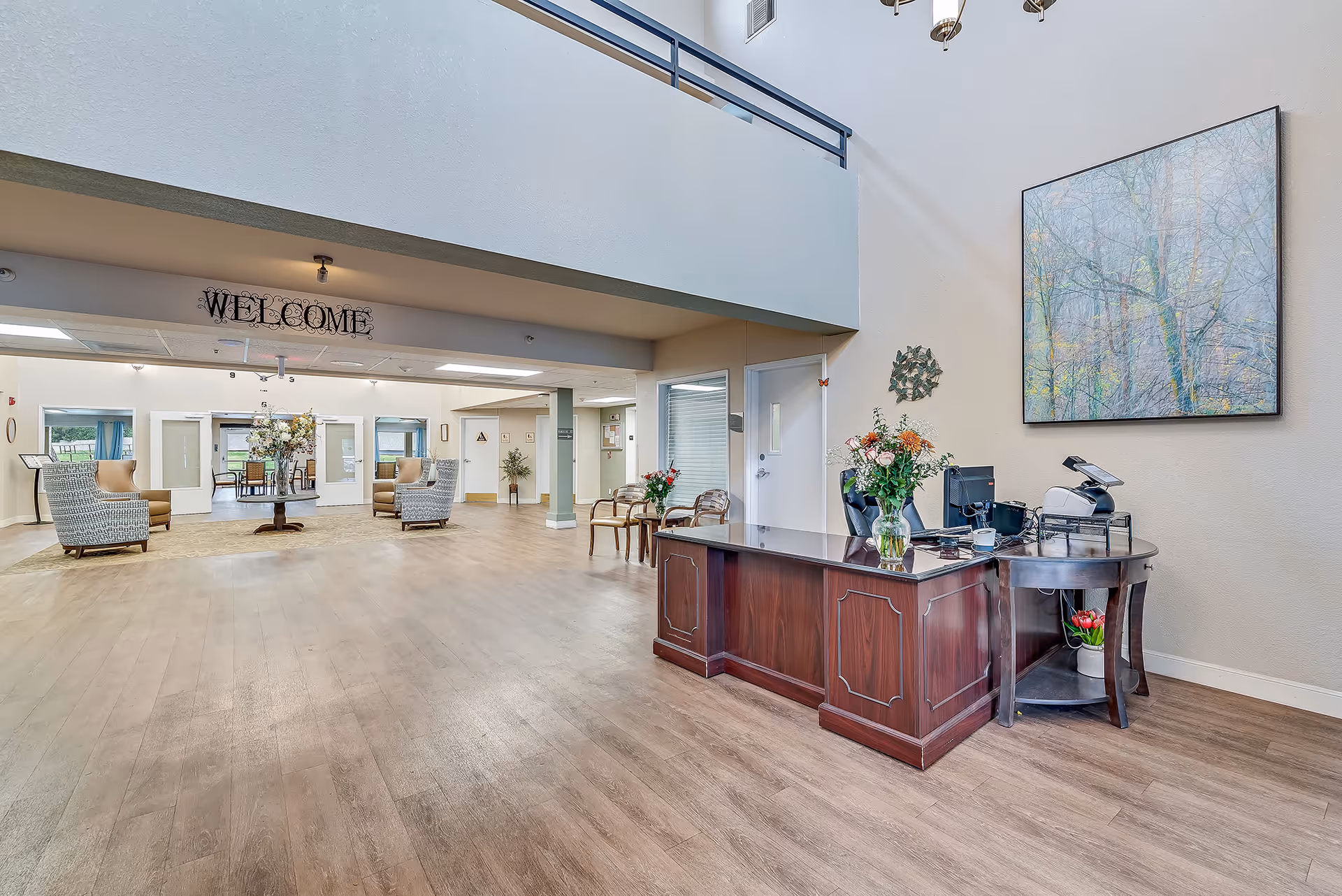 Spacious senior living facility lobby with a wooden reception desk adorned with a vase of flowers, two chairs with a small table and flowers beside them, and a large painting of a forest on the wall. The area has high ceilings, wood flooring, and a welcoming seating area with armchairs and a round table with flowers. A 'WELCOME' sign hangs above the entrance to another room.
