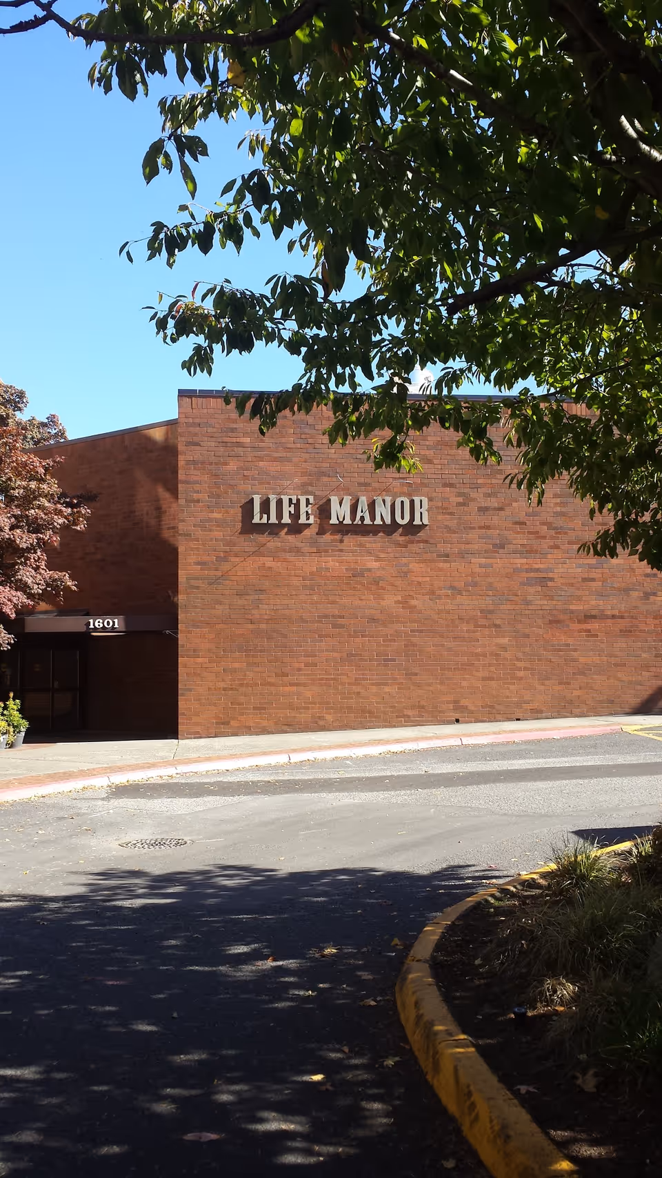 Exterior view of a brick building with the sign 'LIFE MANOR' mounted on the wall. The building entrance is partially visible with the number 1601 above the door. Trees with green leaves frame the top and left side of the image, and a curved driveway with a yellow curb is in the foreground.
