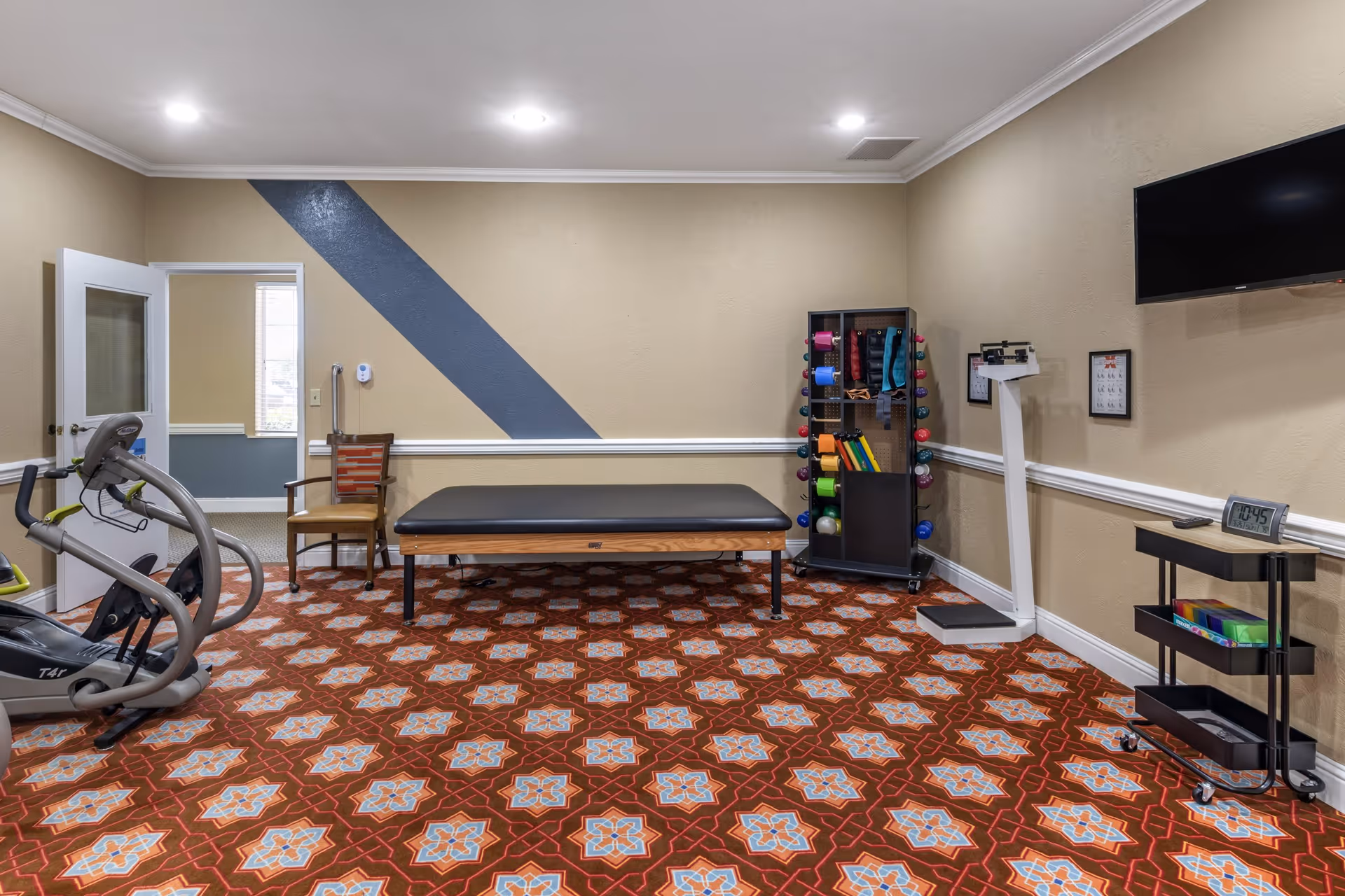 A fitness or physical therapy room with patterned carpet, an exercise bike, a treatment table, a chair, a rack holding various exercise equipment like dumbbells and resistance bands, a scale, a wall-mounted TV, and a small cart with supplies. The walls are beige with white trim and a blue diagonal stripe on one wall.