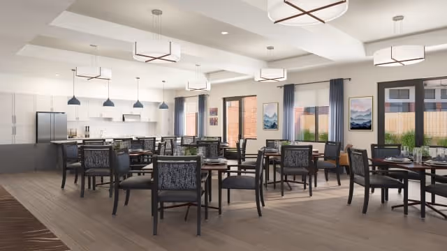 A spacious dining room with multiple tables and chairs arranged neatly. The room features large windows with blue curtains, modern pendant lights, and a kitchen area with stainless steel appliances and white cabinetry in the background. The walls are decorated with framed landscape artwork.