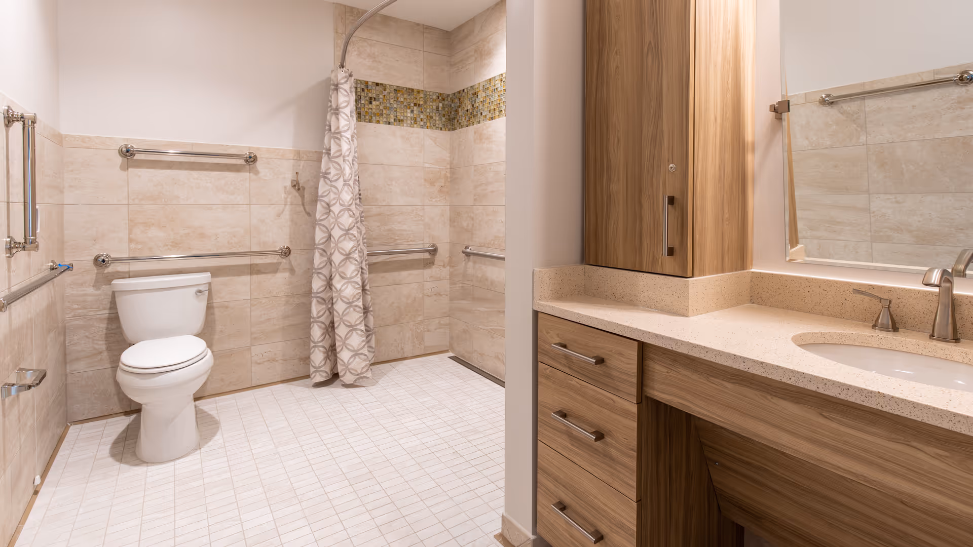 A spacious, modern bathroom with beige tiled walls and floor. The bathroom features a white toilet, a walk-in shower with a patterned curtain, multiple stainless steel grab bars for accessibility, and a wooden vanity with a beige countertop and a sink. A large mirror is mounted above the vanity.