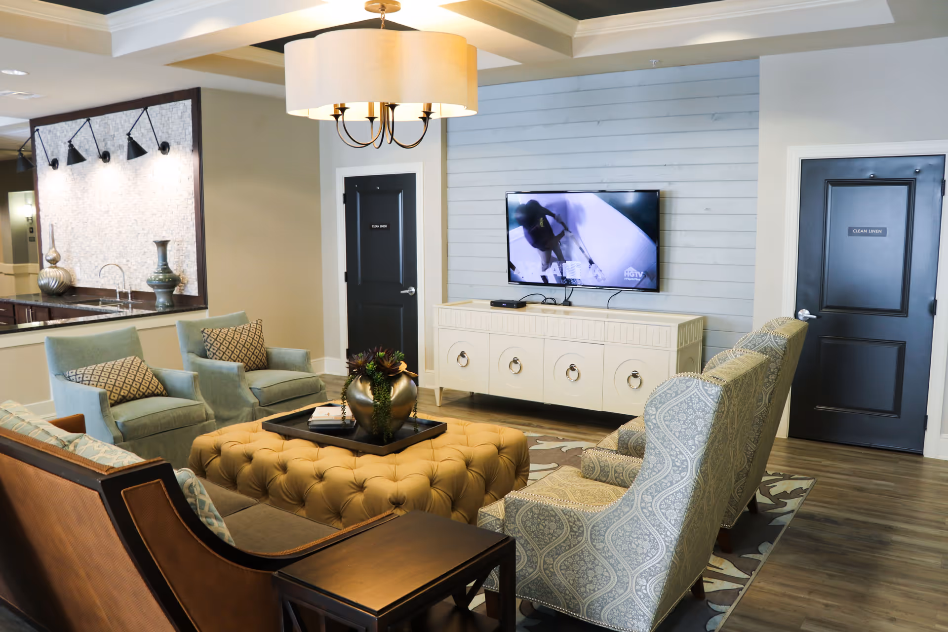A cozy living room area in a senior living facility featuring a large tufted ottoman with a decorative plant centerpiece, two patterned armchairs, two light blue armchairs with patterned pillows, a brown sofa, and a white TV console with a flat-screen TV mounted on a light blue shiplap wall. Two black doors labeled 'CLEAN LINEN' are visible, along with a kitchen counter and decorative vases in the background. A modern chandelier hangs from the ceiling.