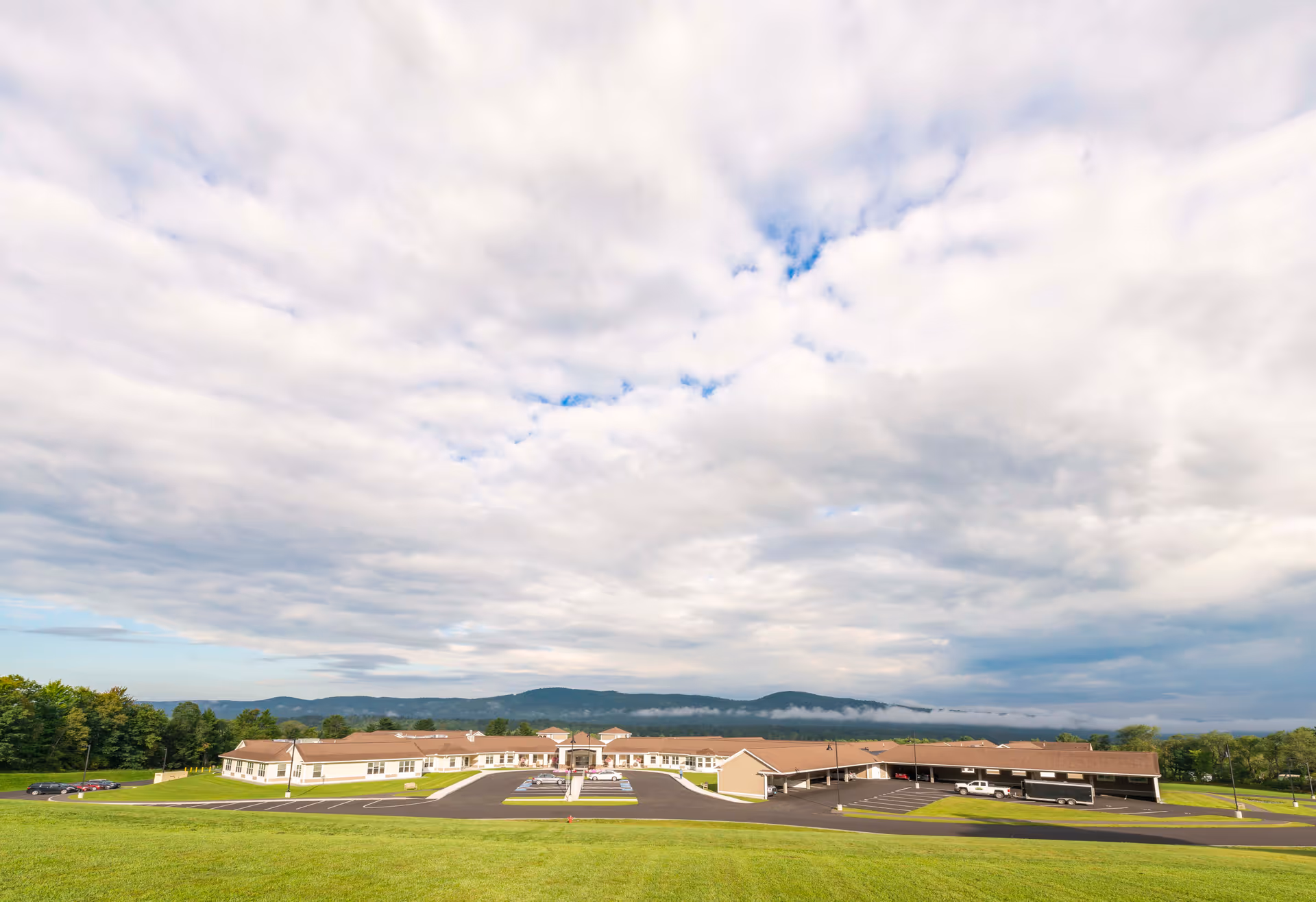 Wide exterior view of a single-story senior living facility named Summit by Morrison, surrounded by green grass and trees with mountains in the background under a cloudy sky.