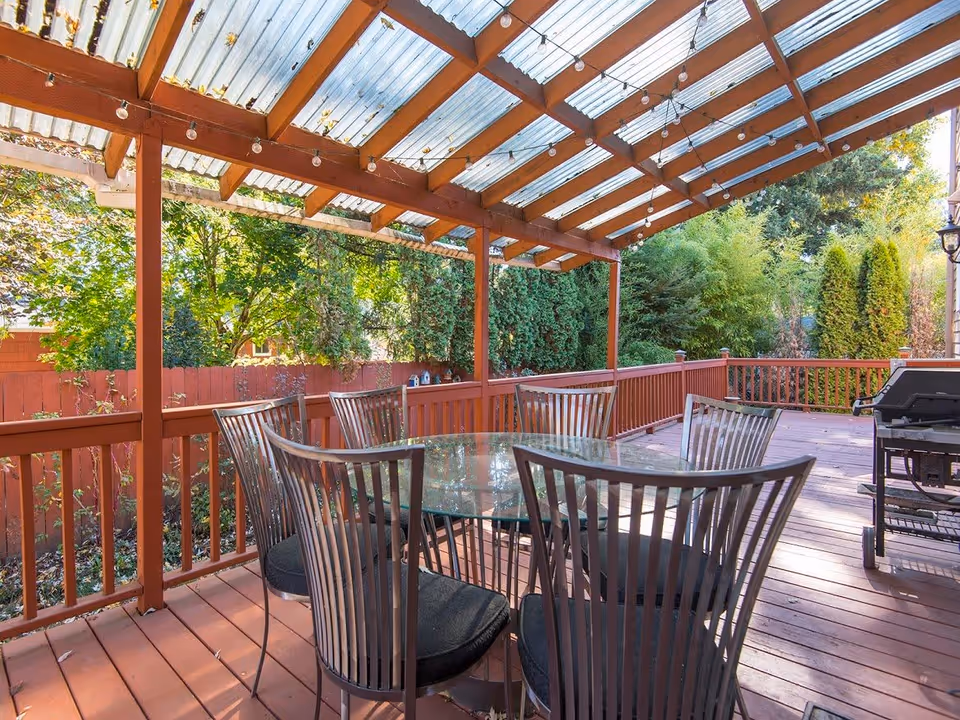 Outdoor covered patio area with a glass-top table surrounded by six metal chairs with black cushions. The patio has a wooden floor and railing painted in reddish-brown. There are string lights hanging from the transparent corrugated roof. Green trees and shrubs are visible beyond the patio fence.
