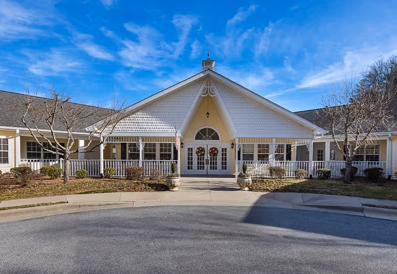 Front exterior view of Carillon Assisted Living of Hendersonville building with a peaked roof, white railings, two wreaths on double doors, and leafless trees on either side under a blue sky with some clouds.