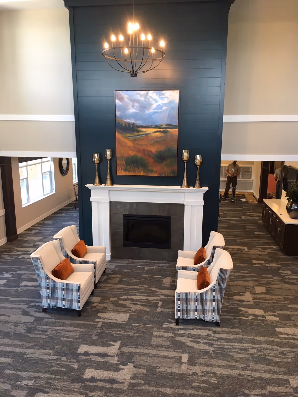 Lounge area with four armchairs arranged around a fireplace beneath a large landscape painting and chandelier.