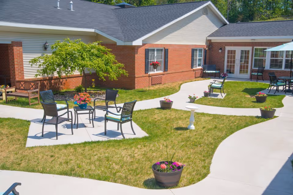 Outdoor courtyard area at Brookridge Heights featuring a paved walkway, several black metal chairs with cushions arranged around small tables, potted flowers, a birdbath, and a bench near a small tree. The building has red brick and beige siding with windows and glass doors leading inside.
