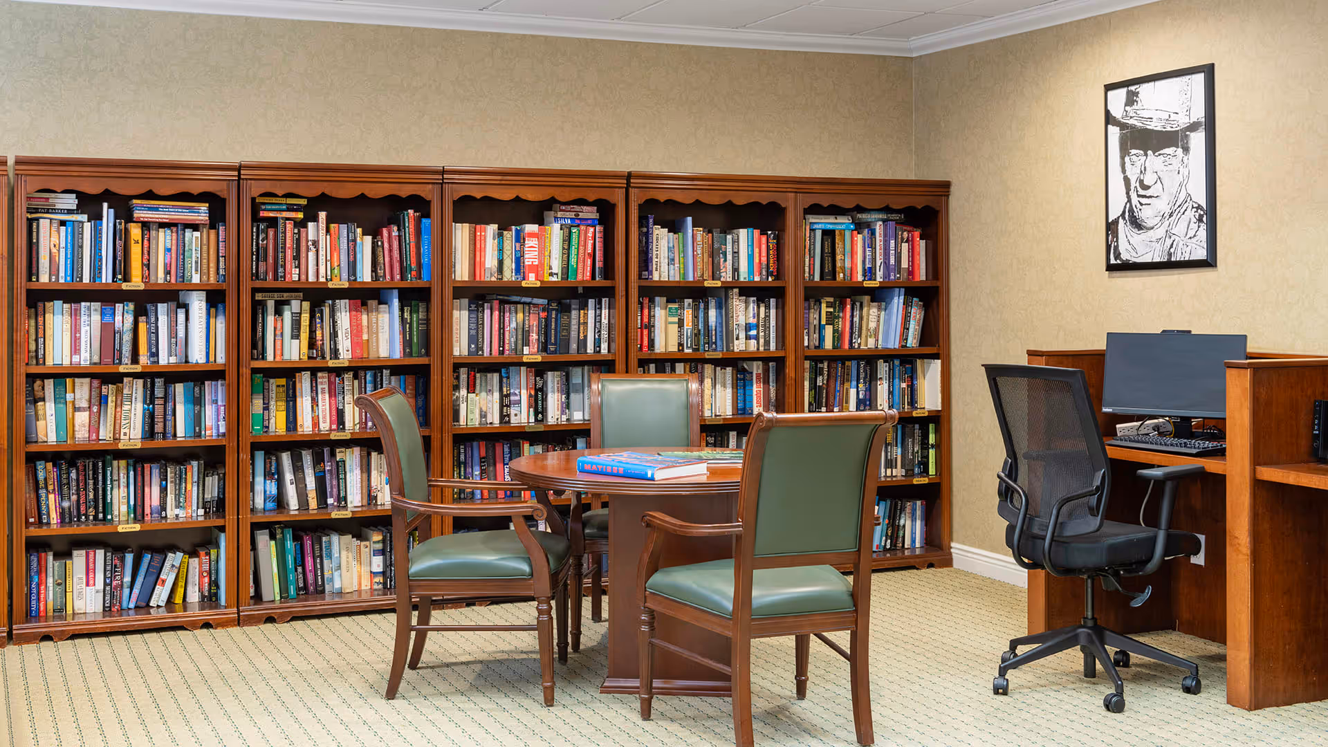 A cozy library room with wooden bookshelves filled with books along the back wall. In front of the shelves, there is a round wooden table with three green cushioned chairs around it. To the right, there is a wooden computer desk with a black mesh office chair and a computer monitor. A black and white portrait hangs on the beige wall above the computer desk.