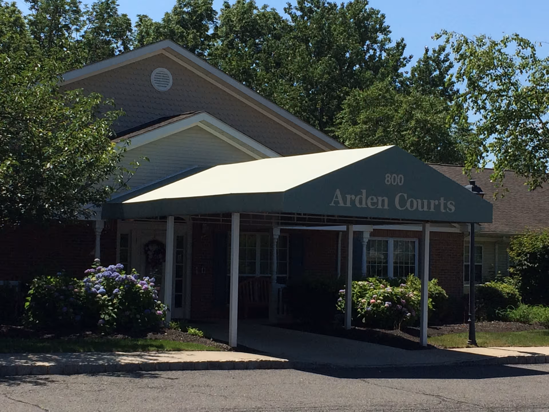 Exterior view of Arden Courts building entrance with a covered awning displaying the name 'Arden Courts' and the number 800. The building is surrounded by greenery and flowering bushes under a clear blue sky.
