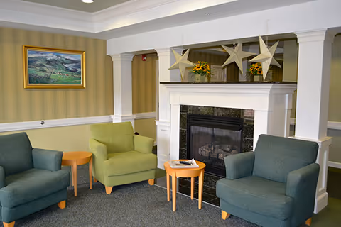 A cozy senior living community lounge area with three armchairs in green and blue shades arranged around two small wooden tables. There is a white fireplace with a black marble surround, decorated with three large star ornaments and two flower arrangements on the mantel. A landscape painting hangs on the wall with striped wallpaper in the background.