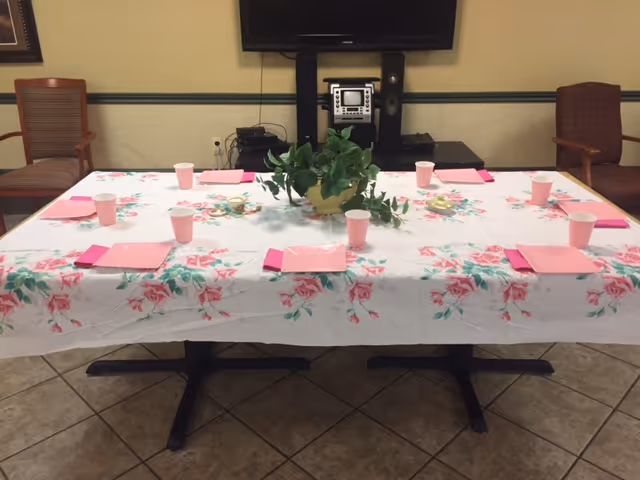 A long table covered with a floral tablecloth set with pink napkins and cups and a potted plant centerpiece in a communal dining area with chairs and a wall-mounted TV.