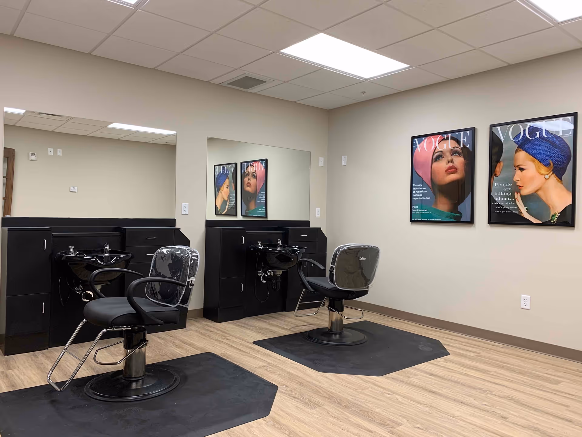Interior view of a salon area in a retirement community featuring two black salon chairs with footrests in front of black washbasins and cabinetry. The walls are beige with two framed Vogue magazine covers hanging on the right wall. The floor is light wood, and the ceiling has white tiles with fluorescent lighting.