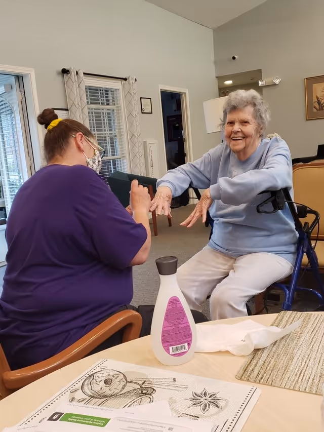 An elderly woman sitting on a walker chair smiling and extending her hands towards a caregiver wearing a purple shirt and a face mask, who is holding the woman's hands. They are in a well-lit room with a table in the foreground that has a bottle of lotion, a paper towel, and placemats on it.