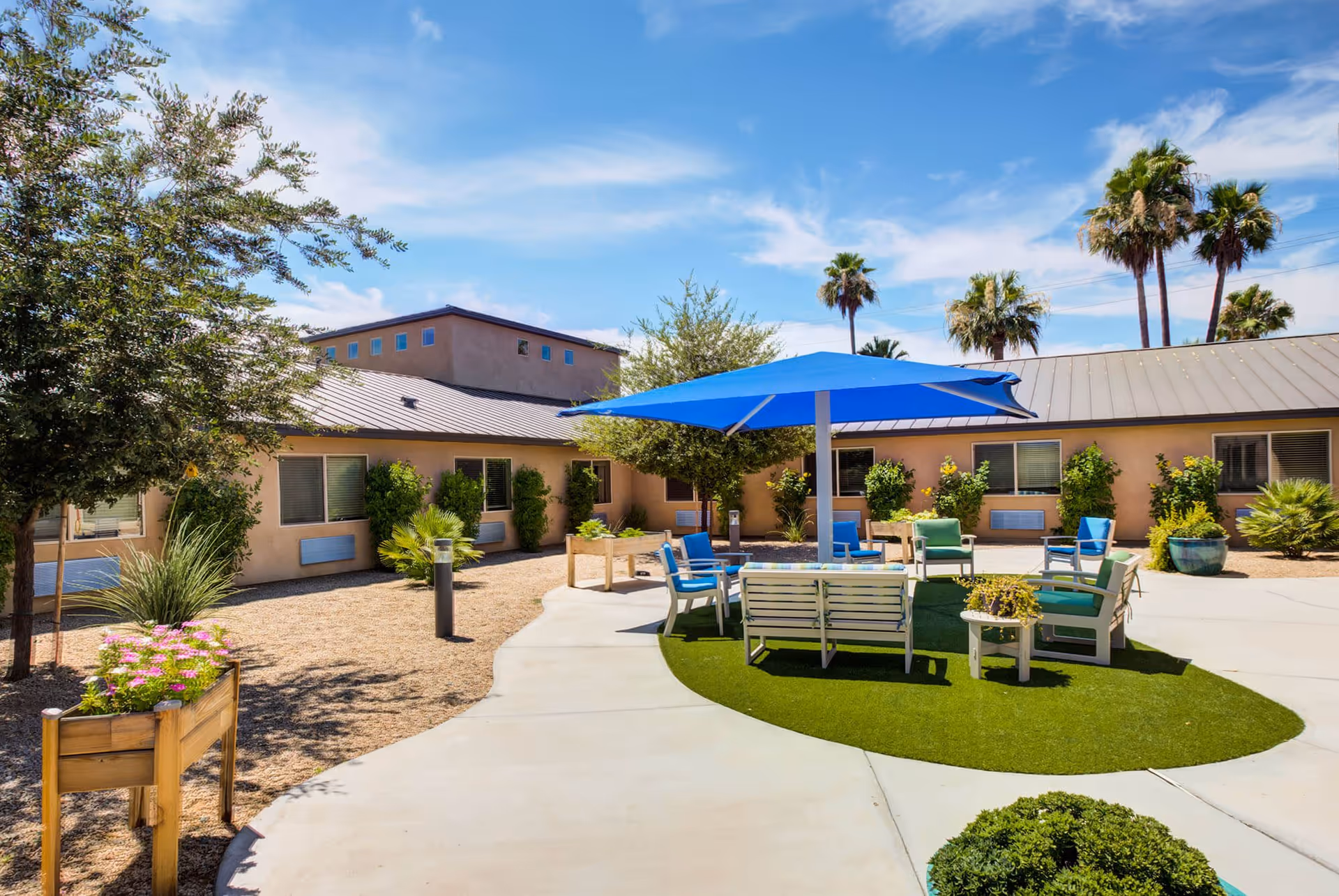 Outdoor courtyard area at Terra Pointe Memory Care featuring a circular seating arrangement with chairs and a bench under a large blue umbrella, surrounded by greenery, potted plants, and a building with multiple windows under a clear blue sky.
