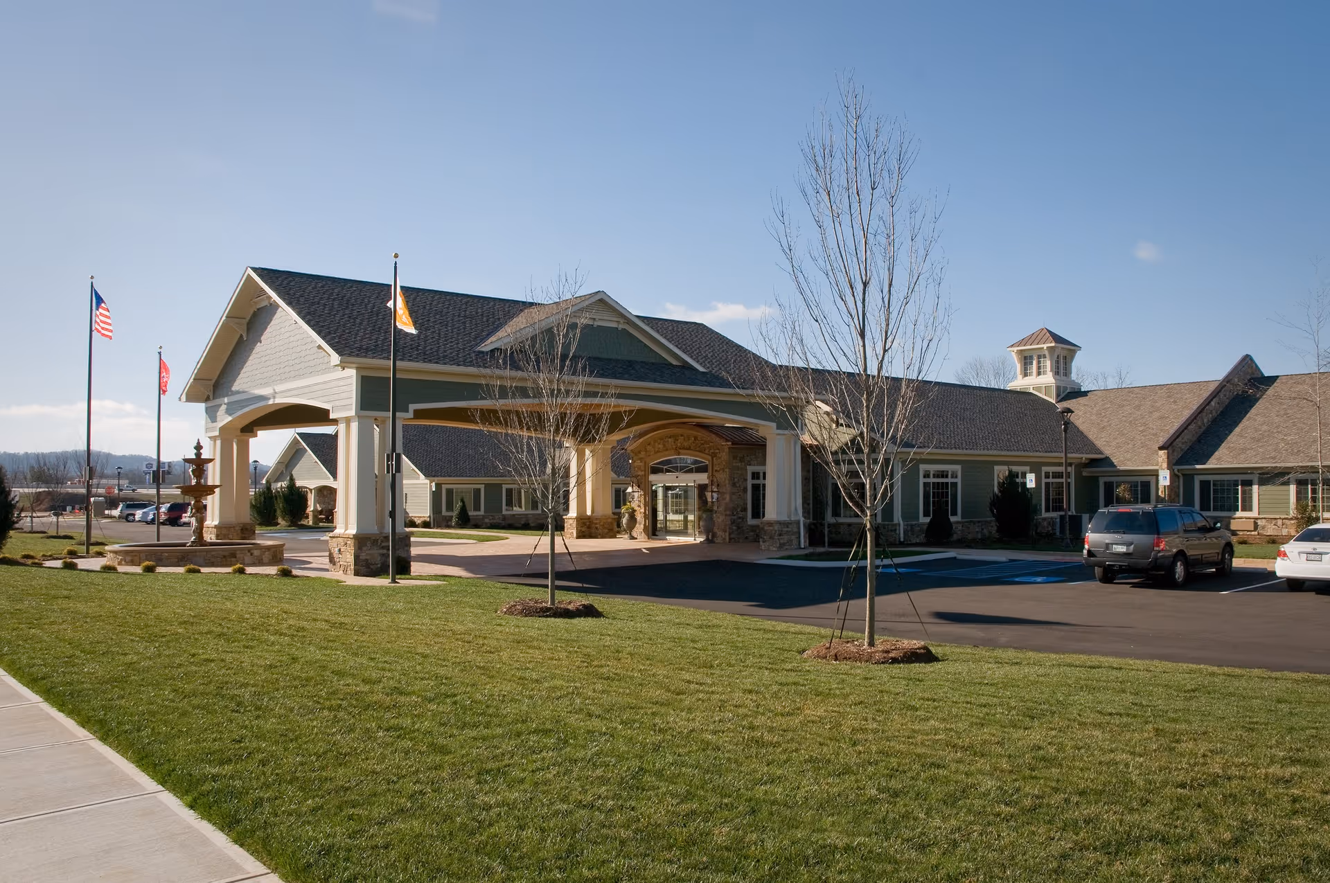 Exterior view of the Life Care Center of Ooltewah building with a covered entrance, several flagpoles, a fountain, parked cars, and a well-maintained lawn under a clear blue sky.